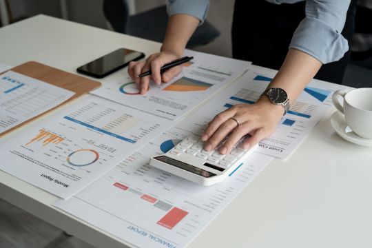 Auf dem Bild sieht man eine Person, die an einem Schreibtisch sitzt und mit Finanzunterlagen arbeitet. Sie benutzt einen Taschenrechner und hält einen Stift in der Hand, während verschiedene Berichte und Diagramme auf dem Tisch verteilt sind. Die Berichte zeigen Balken- und Kreisdiagramme sowie Tabellen. Neben den Unterlagen liegt ein Smartphone, und eine Tasse steht am rechten Bildrand. Die Person trägt eine Uhr am linken Handgelenk und ein hellblaues Hemd. Das Bild vermittelt den Eindruck von analytischer Arbeit oder Finanzplanung.