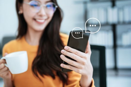 Eine lächelnde Frau in einem gelben Pullover sitzt an einem Schreibtisch und hält ein Smartphone in der Hand. Über dem Smartphone sind Chat-Symbole mit Sprechblasen zu sehen. In ihrer anderen Hand hält sie eine weiße Tasse. Im Hintergrund sind verschwommene Büroregale zu erkennen. Dieses Bild symbolisiert das Konzept der Chatkontrolle, bei dem Nachrichten auf mobilen Geräten überwacht und verwaltet werden.