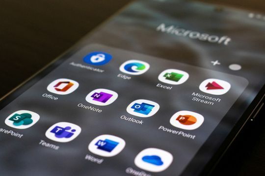 Ein Smartphone-Bildschirm zeigt eine Sammlung von Microsoft-Apps in einem Ordner. Zu den sichtbaren App-Symbolen gehören Authenticator, Edge, Excel, Microsoft Stream, Office, OneNote, Outlook, PowerPoint, SharePoint, Teams, Word und OneDrive. Im Hintergrund ist der Text „Microsoft“ leicht verschwommen zu sehen. Der Bildschirm liegt in einem schrägen Winkel, wodurch die Icons prominent hervorgehoben werden.