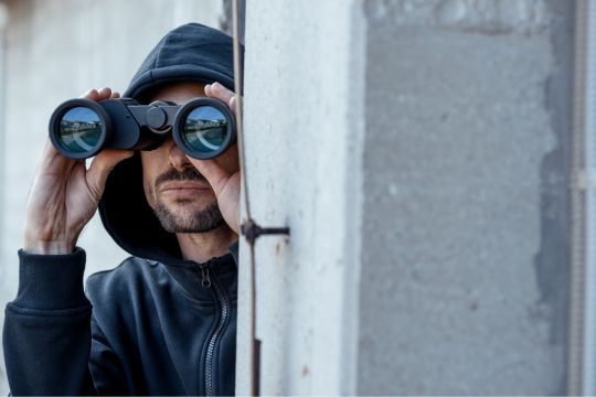 Person mit Kapuze nutzt ein Fernglas, während sie hinter einer Wand hervorschaut