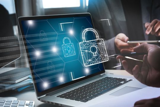 Ein Laptop steht auf einem Schreibtisch, und auf seinem Bildschirm sind digitale Vorhängeschlösser mit vernetzten Linien dargestellt, die für Cyber-Sicherheit und Datenschutz stehen. Im Vordergrund hält eine Hand einen Stift, während eine andere Person im Hintergrund ein Smartphone benutzt. Die Szene vermittelt ein Thema rund um IT-Sicherheit, Datenverschlüsselung und digitale Schutzmaßnahmen.