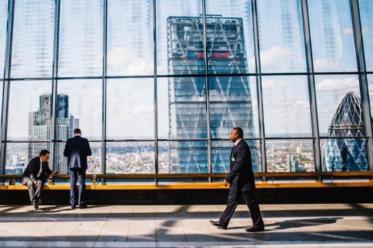 Ein modernes Bürogebäude mit Panoramafenstern und Blick auf die Skyline einer Stadt. Drei Geschäftsleute in Anzügen sind im Bild zu sehen: Einer sitzt und zwei stehen, während sie auf die Stadt hinausschauen. Im Hintergrund sind markante Wolkenkratzer zu erkennen. Das Bild symbolisiert das "kleine Konzernprivileg" und zeigt die Exklusivität und die Vorteile, die mit der Arbeit in einem großen Unternehmen verbunden sind.