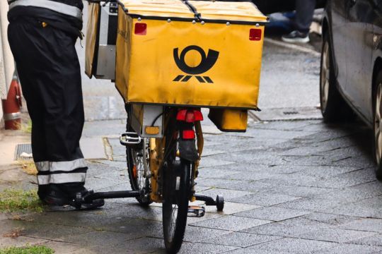 Ein gelbes Postfahrrad steht auf einer nassen Straße. Auf der großen gelben Transportbox des Fahrrads ist das Logo der Post zu sehen. Neben dem Fahrrad steht eine Person in einer dunklen Arbeitsuniform, von der nur die Beine sichtbar sind. Die Szenerie wirkt wie ein typischer Zustellmoment bei Regenwetter oder nach einem Regenschauer.